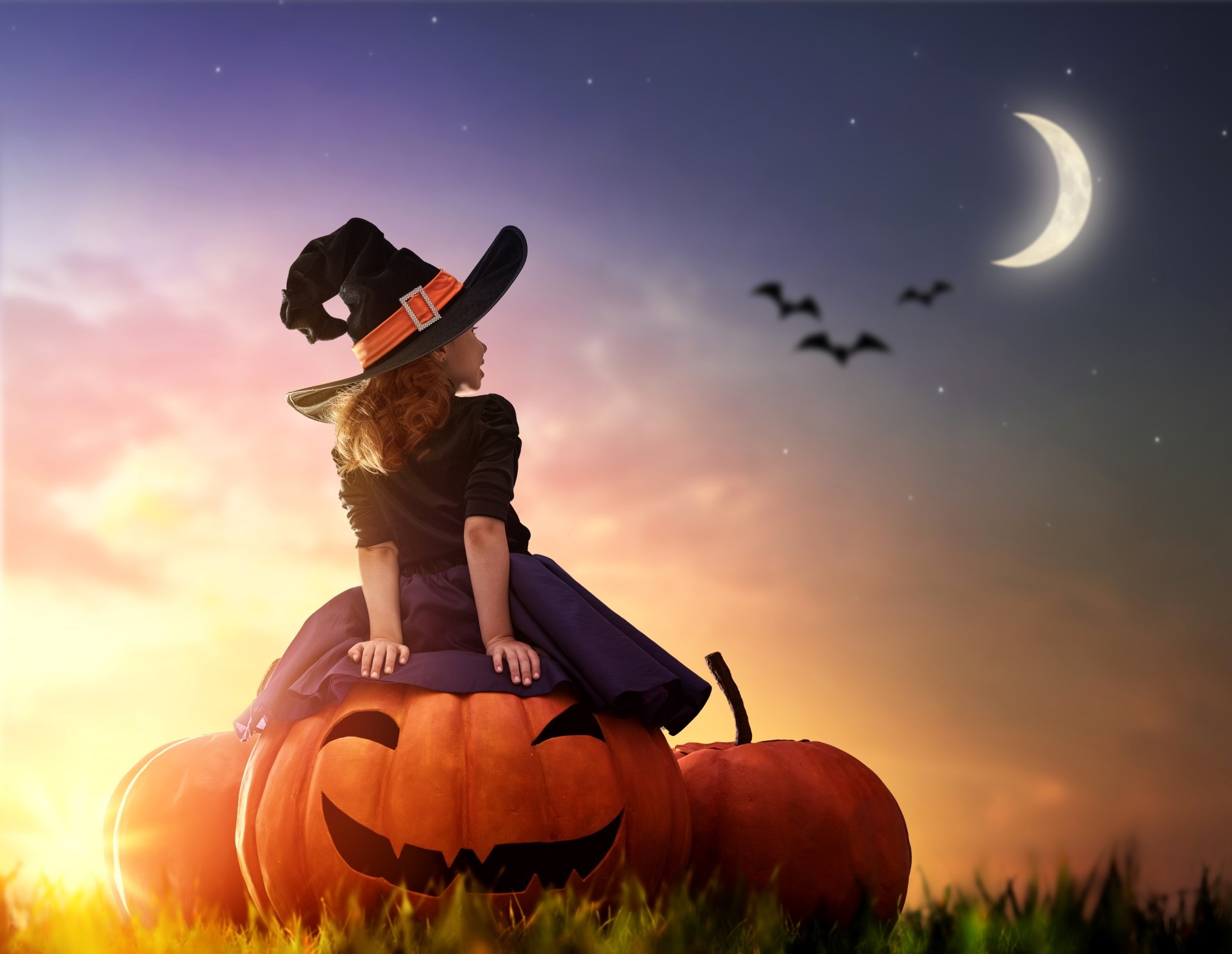 A child wearing a witch hat sits on a large pumpkin under a crescent moon with bats flying, captured in a 4K Ultra HD Halloween-themed desktop wallpaper.