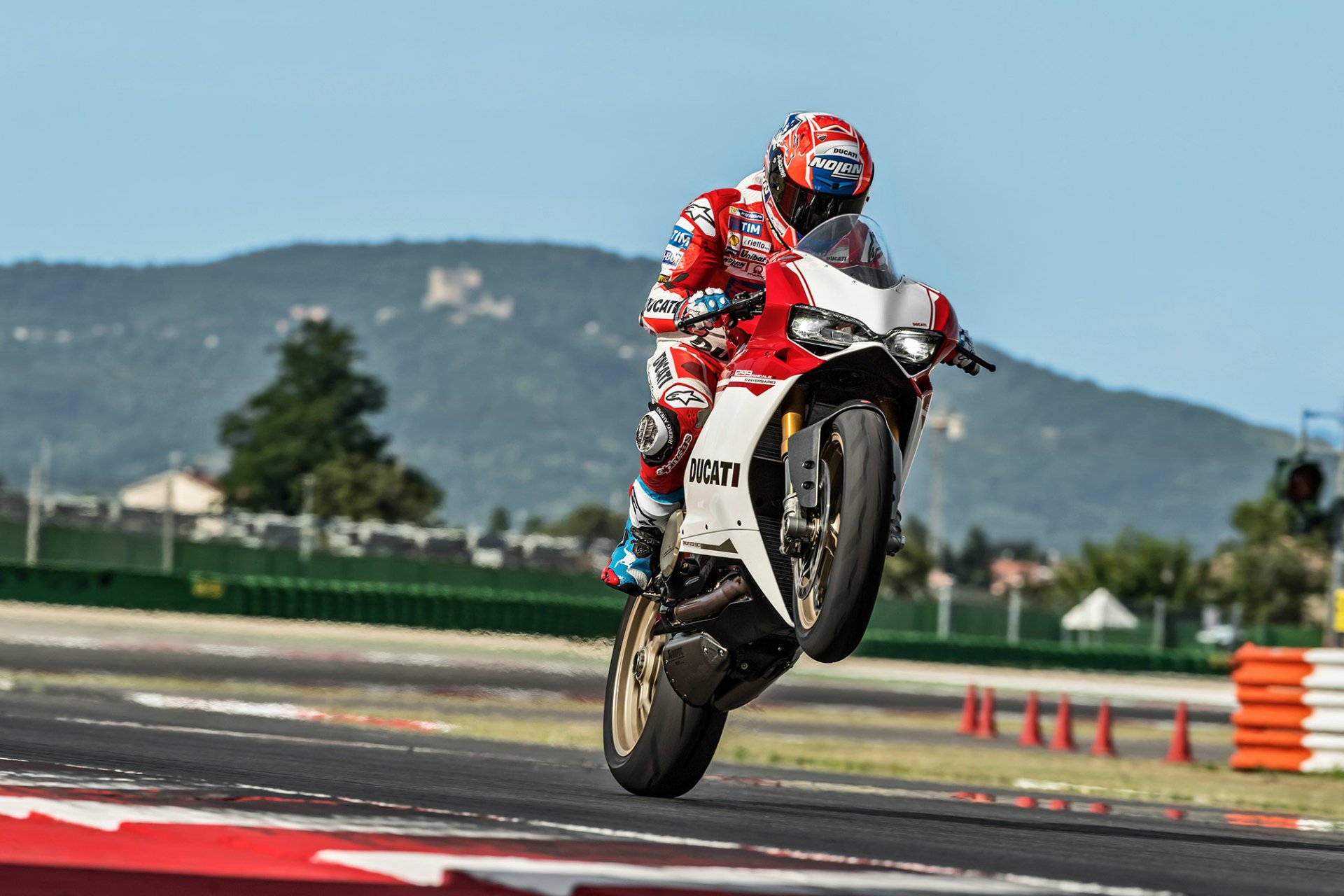 HD desktop wallpaper of a Ducati 1299 motorcycle performing a wheelie on a racetrack, rider in red leathers and helmet with hills in the background.