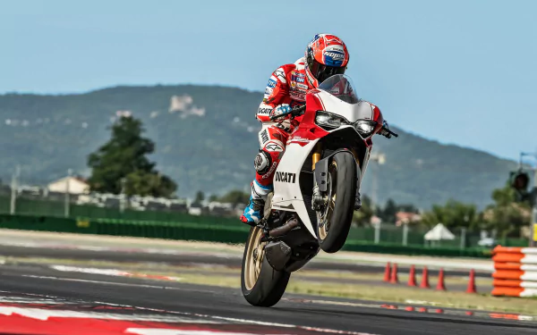 HD desktop wallpaper of a Ducati 1299 motorcycle performing a wheelie on a racetrack, rider in red leathers and helmet with hills in the background.