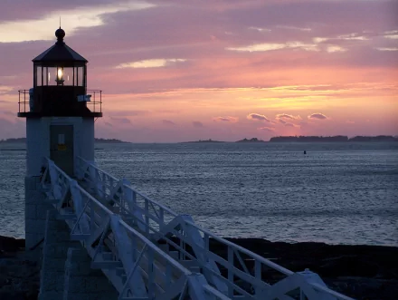 HD desktop wallpaper of a man-made lighthouse with an illuminated lantern, overlooking a tranquil ocean at sunset. The sky is painted in shades of pink and orange, creating a serene and picturesque background.