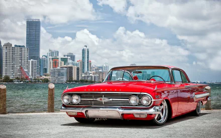Bright red Chevrolet Impala hot rod parked by the waterfront with a city skyline under a partly cloudy sky, captured in HD for a striking desktop wallpaper.