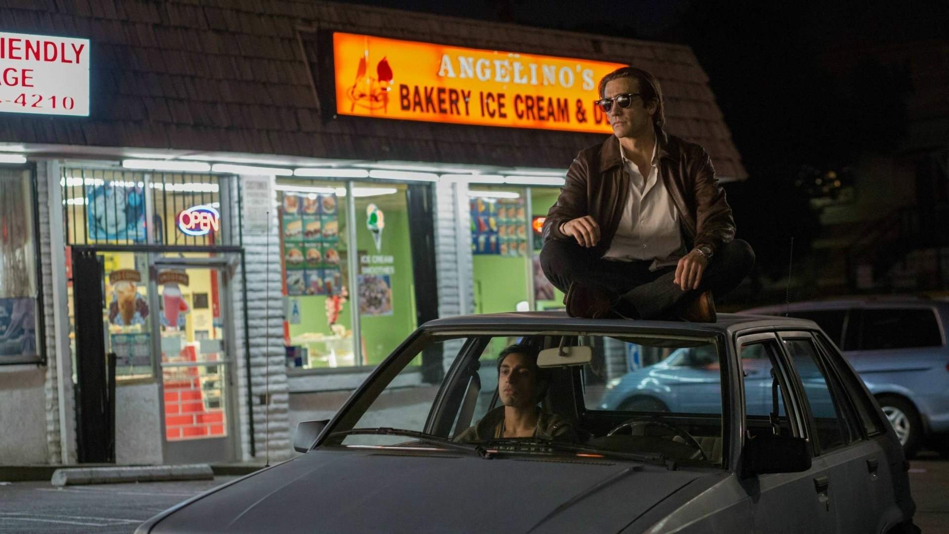 HD desktop wallpaper featuring a night scene from the movie Nightcrawler (2014) with a man sitting on a car roof outside Angelino’s Bakery Ice Cream & Deli.