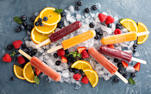 Vibrant 4K wallpaper featuring orange, blueberry, and raspberry popsicles with fresh berries, orange slices, and ice cubes on a textured surface.