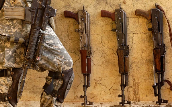 A soldier stands near several AK-47 and AKM assault rifles against a textured wall, showcasing the intricate details of these man-made weapons in a high-definition wallpaper.