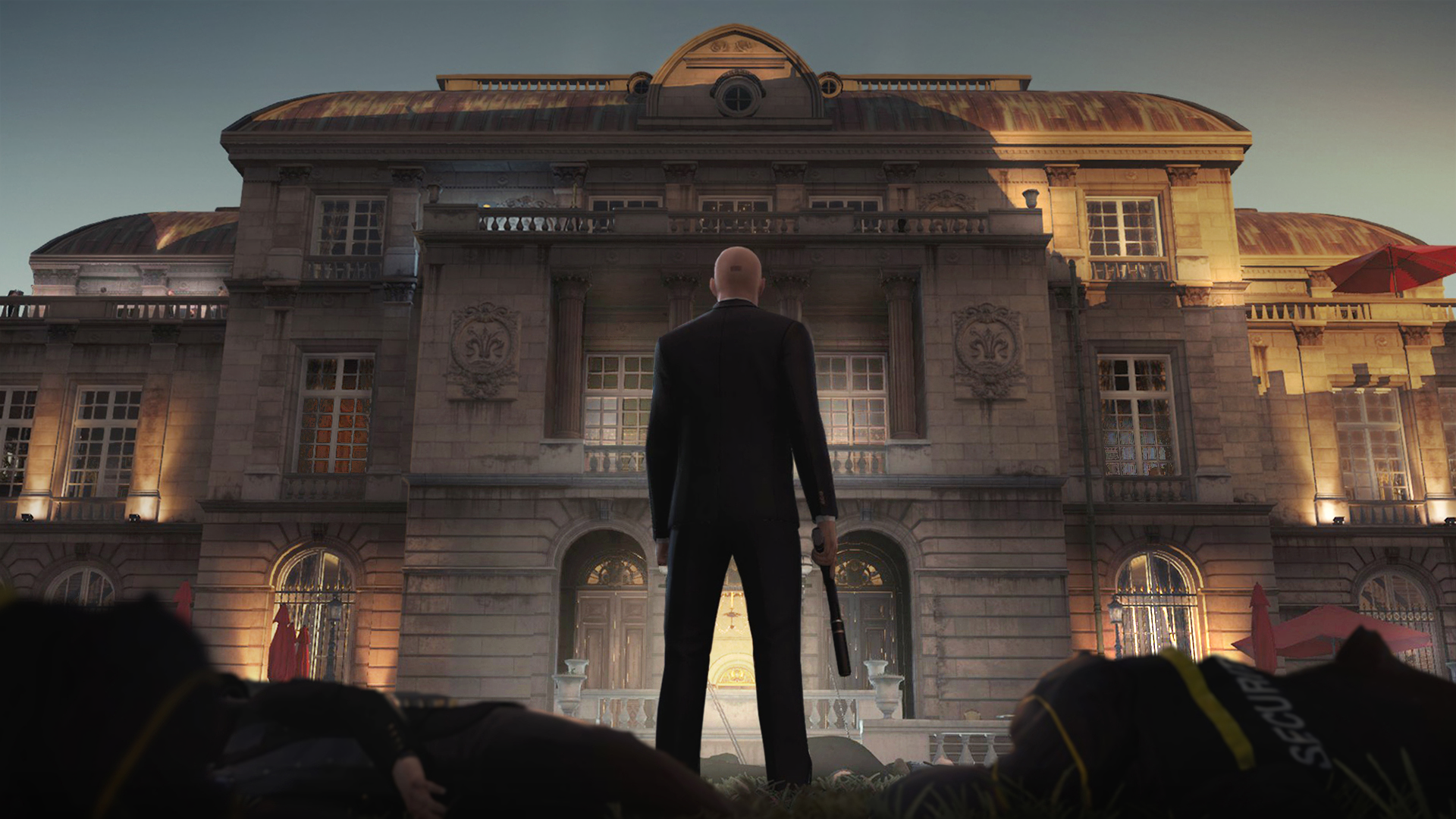 HD PC desktop wallpaper featuring a scene from the video game Hitman (2016), showing Agent 47 standing in front of a grand, illuminated building at dusk.