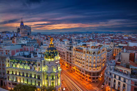 Evening cityscape of Madrid, Spain, showcasing illuminated streets and historic buildings under a dramatic sky in this HD desktop wallpaper.