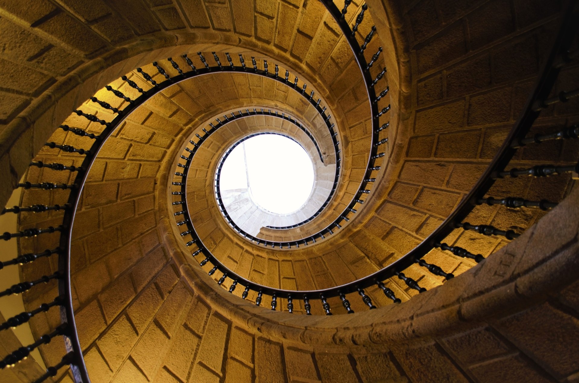 Spiral stone staircase viewed from below, man-made stairs curling into a bright circular skylight — 2K Quad HD PC desktop wallpaper/background.