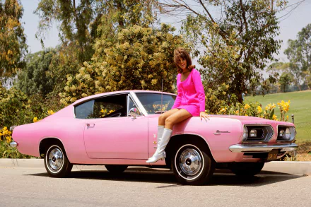 A woman in a pink dress and white boots sits on the hood of a pink Plymouth car, surrounded by greenery in this HD PC desktop wallpaper featuring girls and cars.