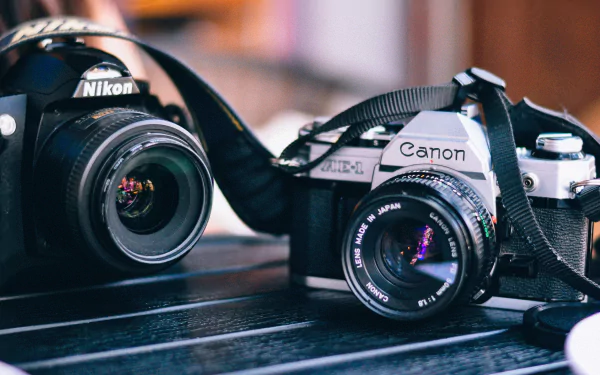 4K Ultra HD PC desktop wallpaper of two man-made cameras: a Nikon and a Canon SLR on a table, lenses catching colorful bokeh.