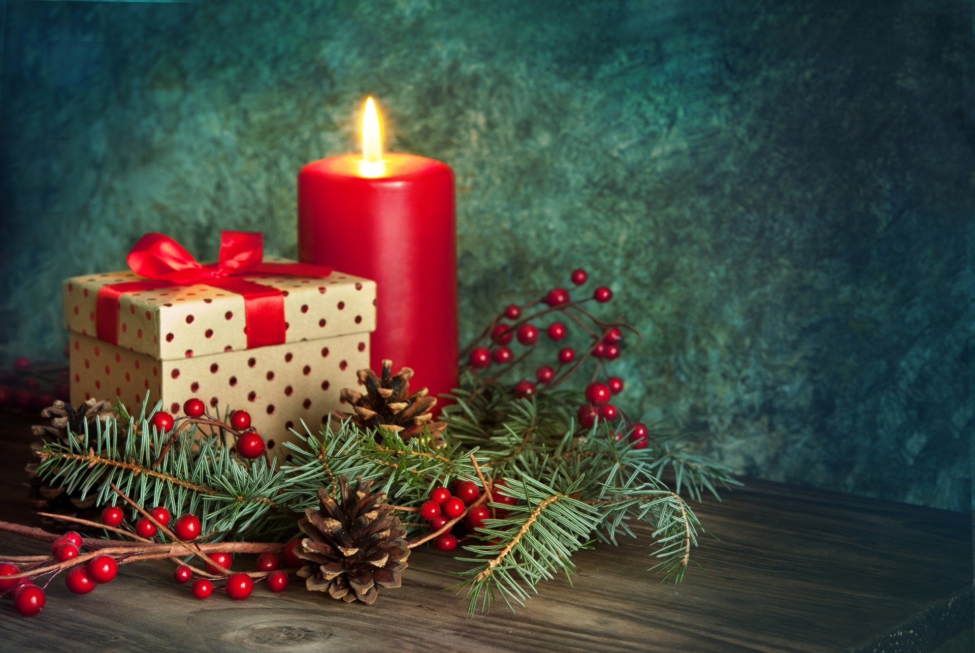 HD desktop wallpaper featuring a festive Christmas scene with a lit red candle, a gift box wrapped in polka dot paper with a red ribbon, pine cones, and evergreen branches with red berries.