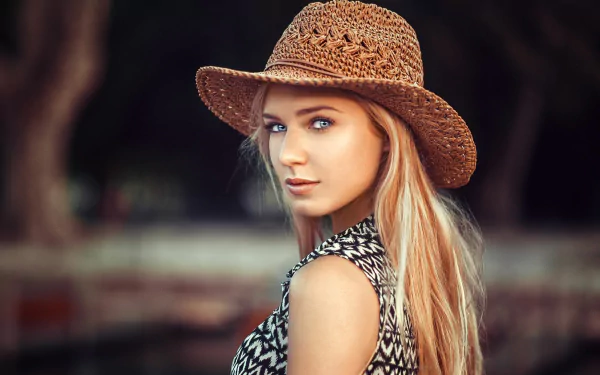 Blonde woman with blue eyes wearing a woven hat, posing outdoors with a blurred natural background. HD desktop wallpaper featuring model Eva Mikulski.