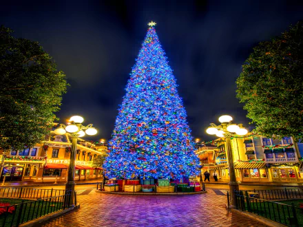 A brightly lit blue Christmas tree stands in the center of a festive town square at night, surrounded by holiday lights and buildings under a dark sky.