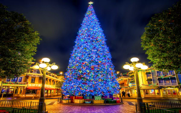 A brightly lit blue Christmas tree stands in the center of a festive town square at night, surrounded by holiday lights and buildings under a dark sky.