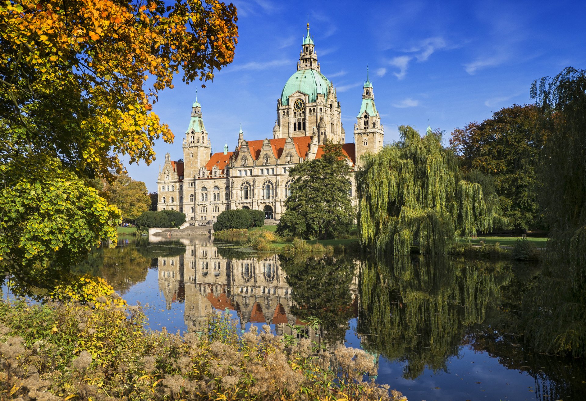 View of Hanover's New City Hall in Germany, showcasing its detailed architecture reflected in a serene body of water, framed by autumn foliage.