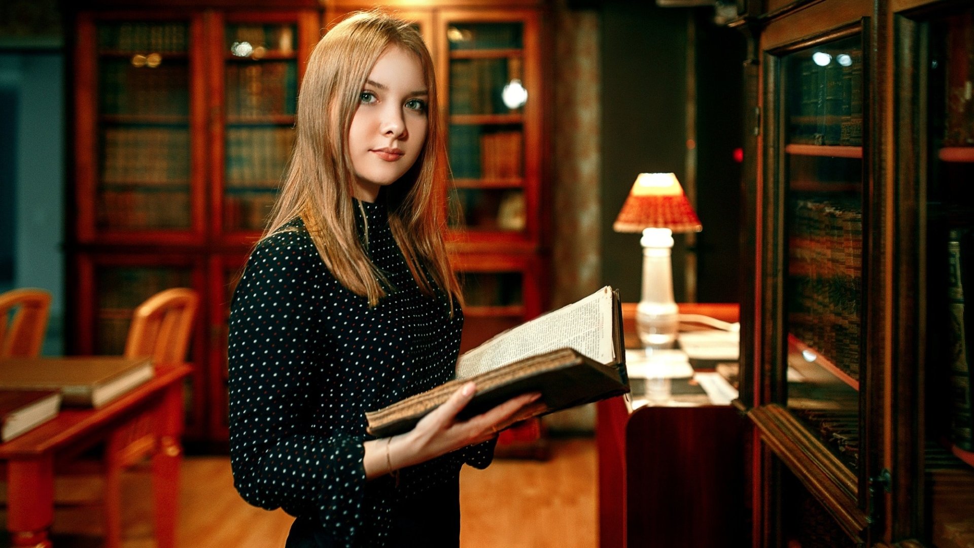 A blonde woman with green eyes holds an open book in a well-lit library, surrounded by wooden bookshelves and a table lamp. HD desktop wallpaper.