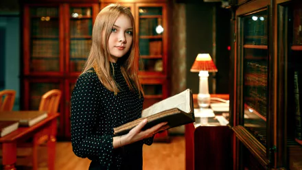 A blonde woman with green eyes holds an open book in a well-lit library, surrounded by wooden bookshelves and a table lamp. HD desktop wallpaper.