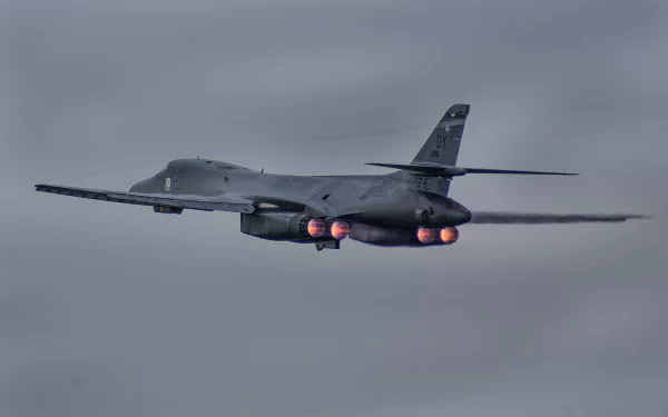 Rockwell B-1 Lancer military bomber aircraft in flight with afterburners engaged against a cloudy sky, shown in HD desktop wallpaper quality.