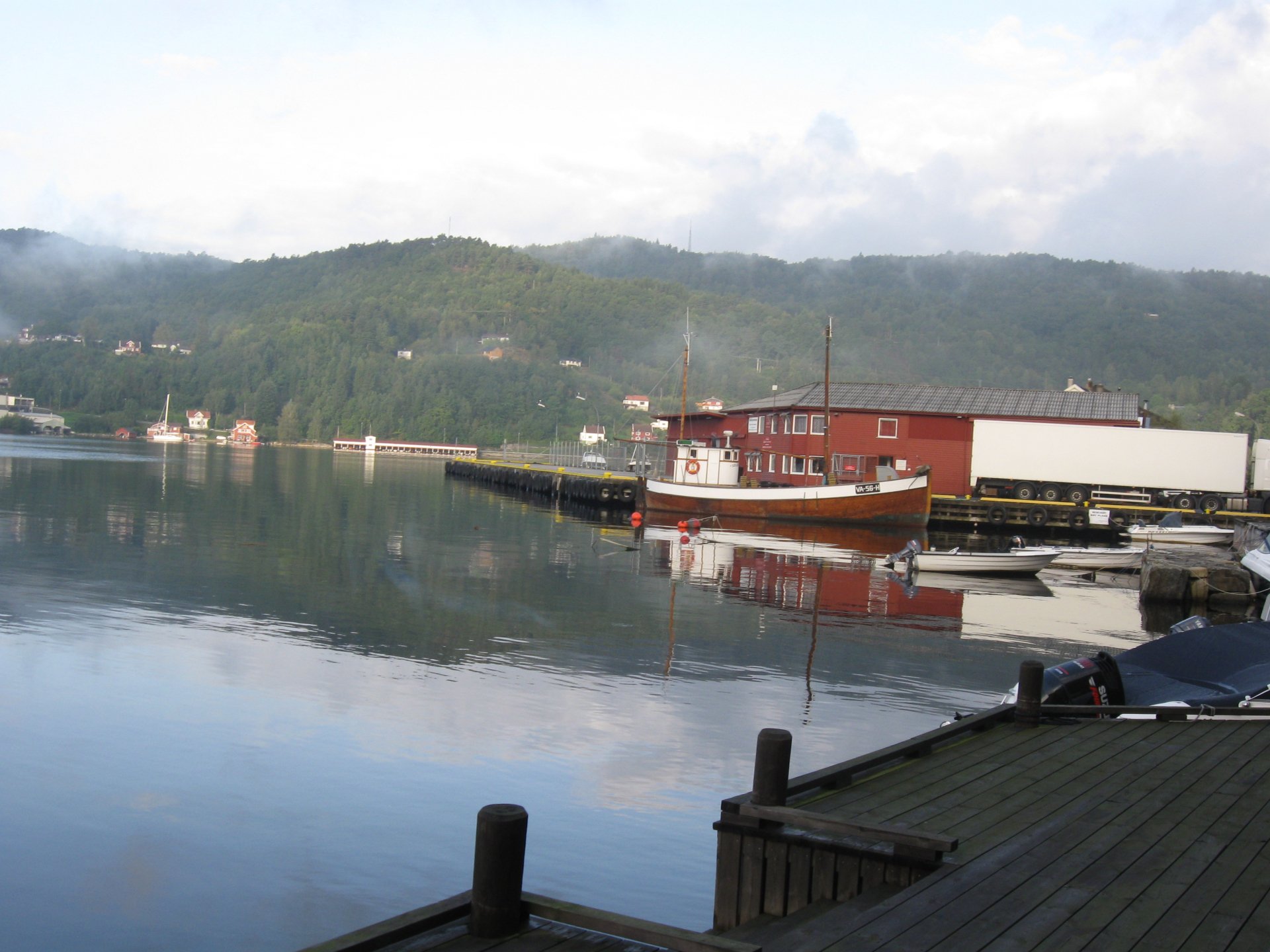 HD desktop wallpaper showcasing a serene man-made harbor with calm waters reflecting a docked boat and lush green hills in the background under a cloudy sky.