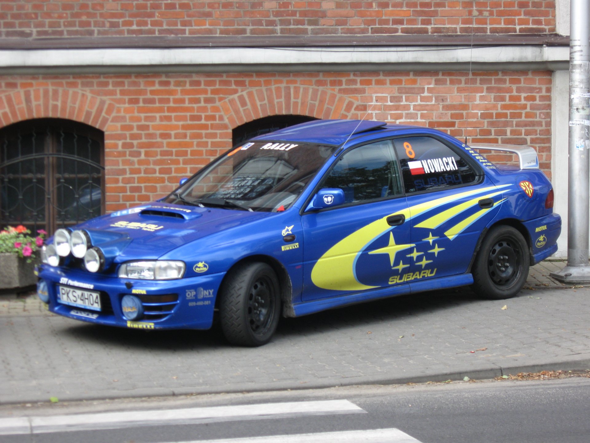 Blue Subaru Impreza rallye vehicle with yellow graphics parked by a brick wall — 2K Quad HD PC desktop wallpaper/background.
