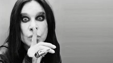 Black-and-white HD PC wallpaper of a heavy metal singer with long dark hair and dramatic eye makeup, holding a finger to lips in a shushing pose against a gray background.