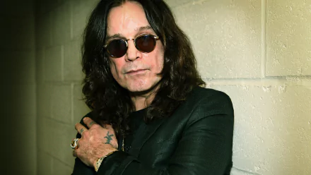 HD PC desktop wallpaper: heavy metal music portrait of a long-haired musician in sunglasses and a black jacket leaning against a pale cinderblock wall.