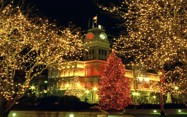 A beautifully lit town square at night with a decorated Christmas tree, festive holiday lights, and a historic building glowing against the dark sky.
