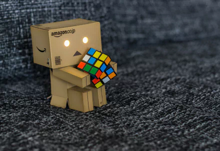 A Danbo figure sitting on a textured surface, holding and examining a colorful Rubik's Cube, captured in HD for a PC desktop wallpaper background.