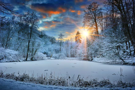 HD desktop wallpaper featuring a serene winter landscape at sunset, with snow-covered trees surrounding a frozen lake, and vibrant orange and blue skies in the background.