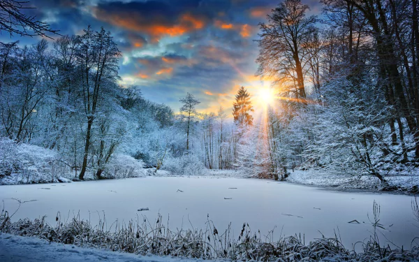 HD desktop wallpaper featuring a serene winter landscape at sunset, with snow-covered trees surrounding a frozen lake, and vibrant orange and blue skies in the background.