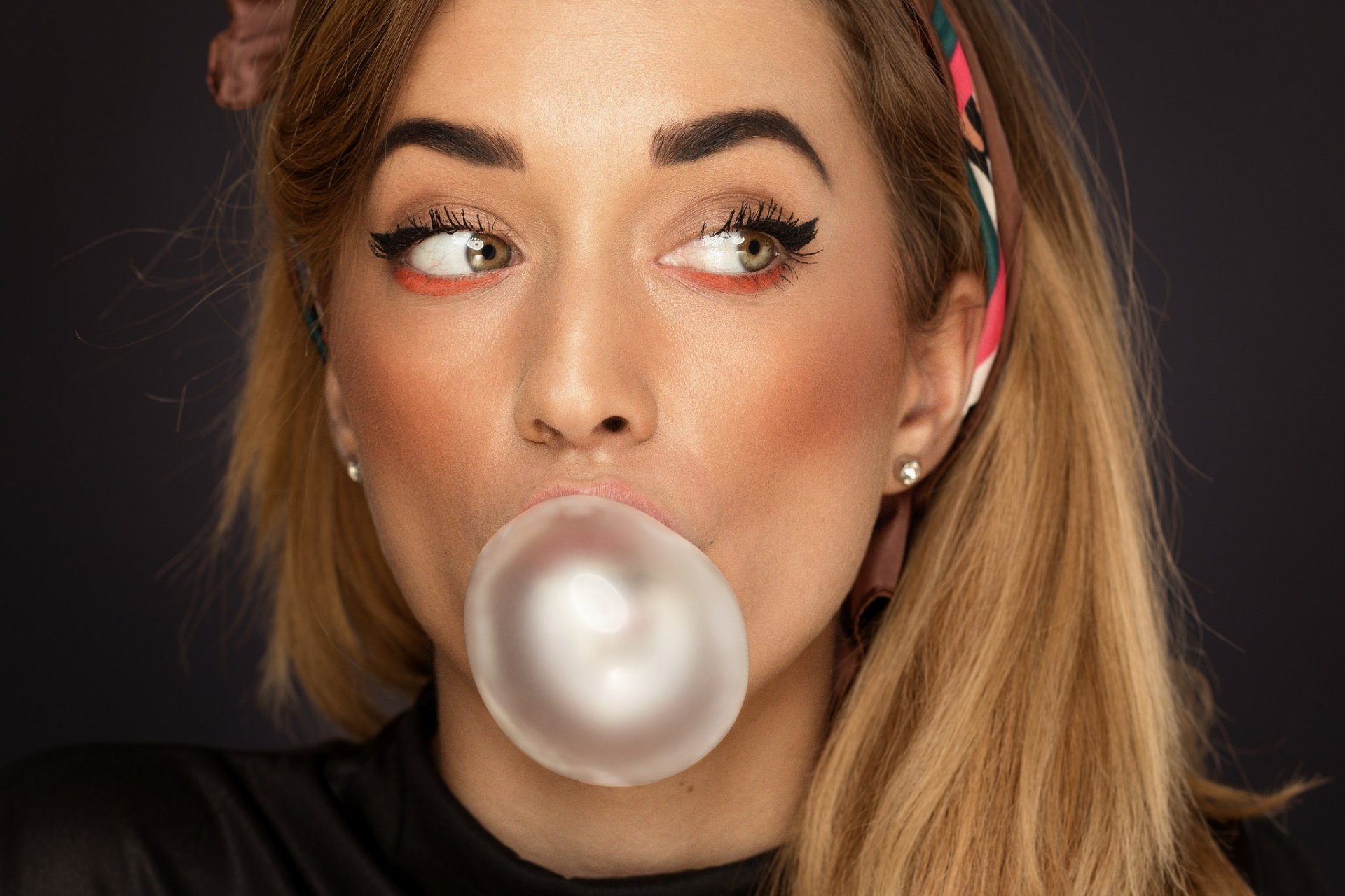 Blonde woman with hazel eyes and flawless makeup blows a bubble with chewing gum, captured in a high-definition close-up model portrait.