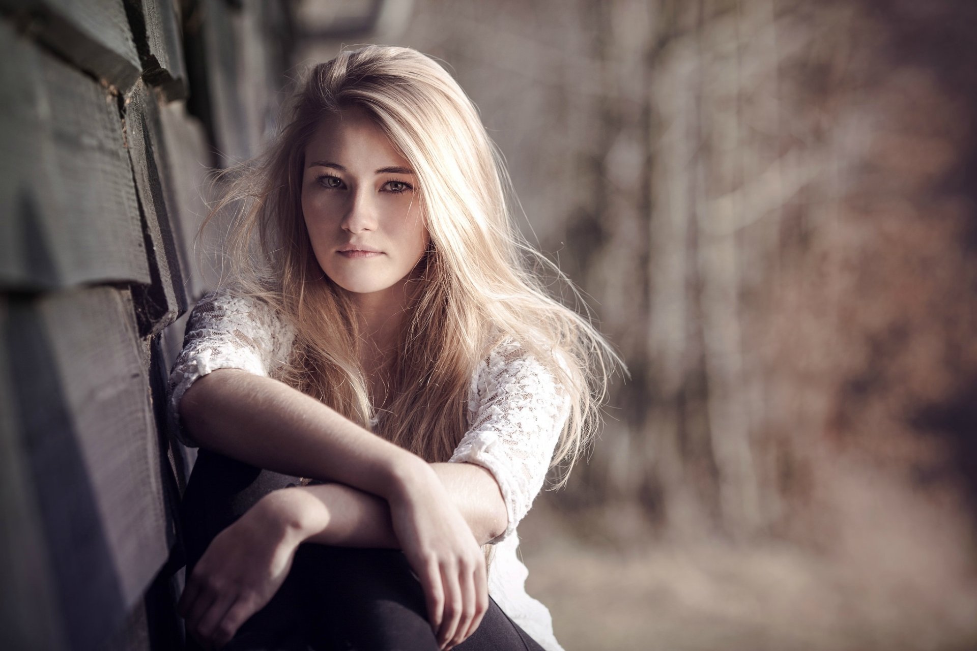 Blonde woman with long hair and hazel eyes sits against a textured wall in soft natural light, captured with shallow depth of field for an HD desktop wallpaper.