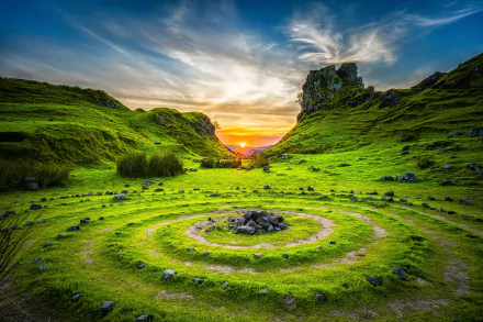 HD desktop wallpaper of a stunning nature landscape at sunset, featuring a vibrant green field with a spiral stone pattern and rocky hills in the background.
