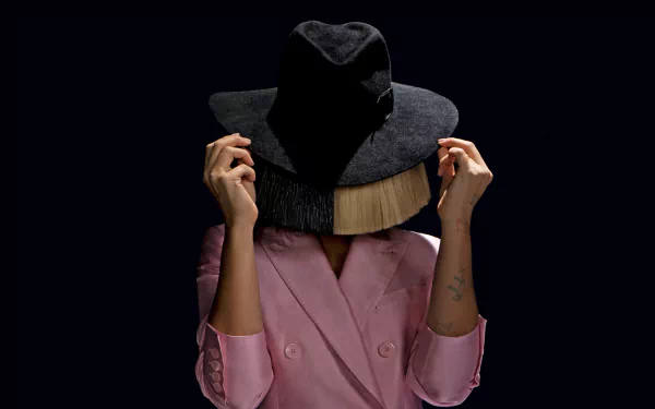 HD desktop wallpaper featuring a person in a pink jacket holding a black hat over their face, evoking the style of Sia.