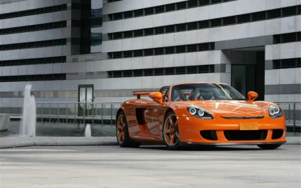 An orange Porsche 911 Carrera parked in an urban setting, showcasing its sleek design against modern architecture, featured as an HD PC desktop wallpaper.