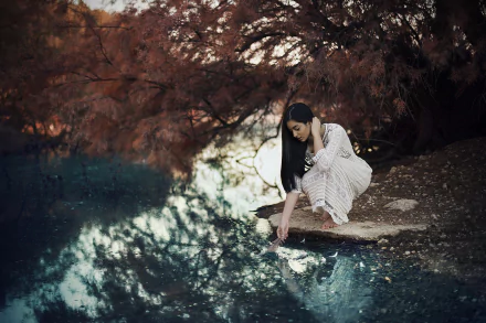A brunette woman in a white dress crouches by a river, touching the water’s reflection amid autumnal trees, creating a serene and moody fall scene.