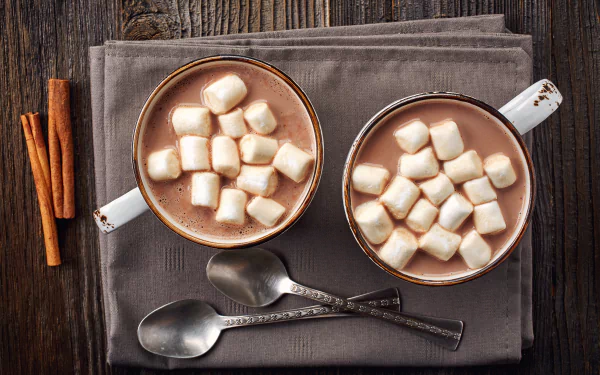 Two cups of hot chocolate topped with marshmallows sit on a cloth next to cinnamon sticks and spoons, captured in 4K Ultra HD for a PC desktop background.