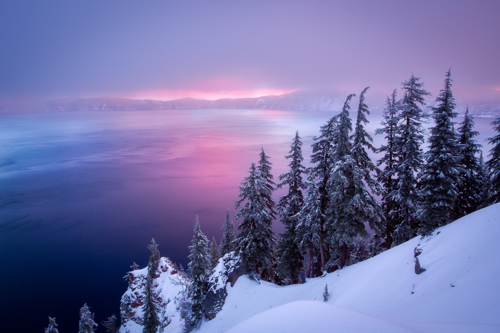 Snow-covered trees overlook the foggy, purple-hued waters of Crater Lake in this serene winter nature scene.