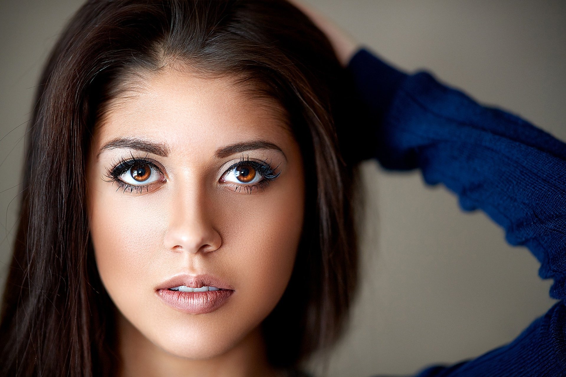 Close-up HD desktop wallpaper of a brunette woman with striking brown eyes and a neutral expression, her face framed by dark hair and a blue sleeve.