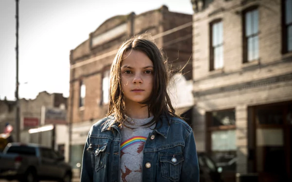 HD desktop wallpaper featuring Dafne Keen as Laura Kinney, also known as X-23, from the movie Logan, standing against a rustic urban backdrop.