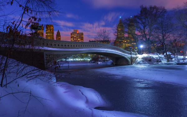 Winter view of Bow Bridge in Central Park, New York, illuminated at dusk with snow-covered banks and city skyline in the background, captured in a HD desktop wallpaper.