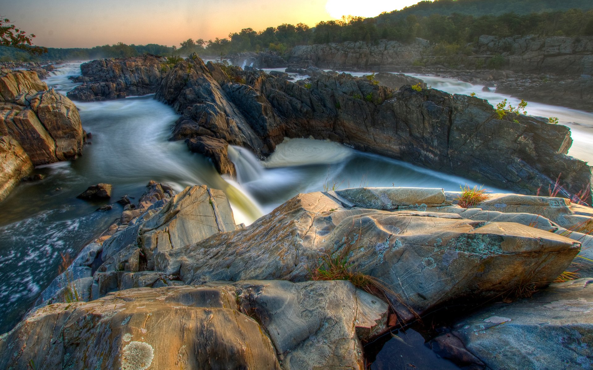 HD PC desktop wallpaper capturing a serene river flowing through rugged rocks in a natural landscape at sunset.