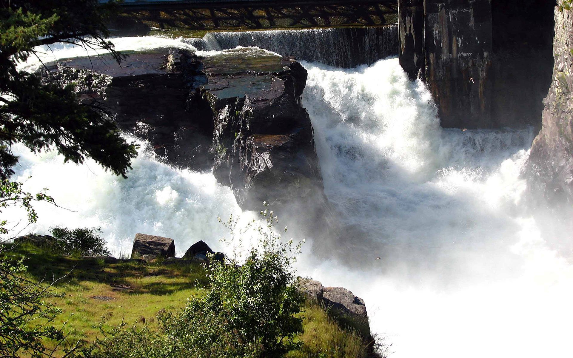 HD PC desktop wallpaper featuring a powerful waterfall cascading over rocks surrounded by lush greenery and a bridge overhead, showcasing the beauty of nature.