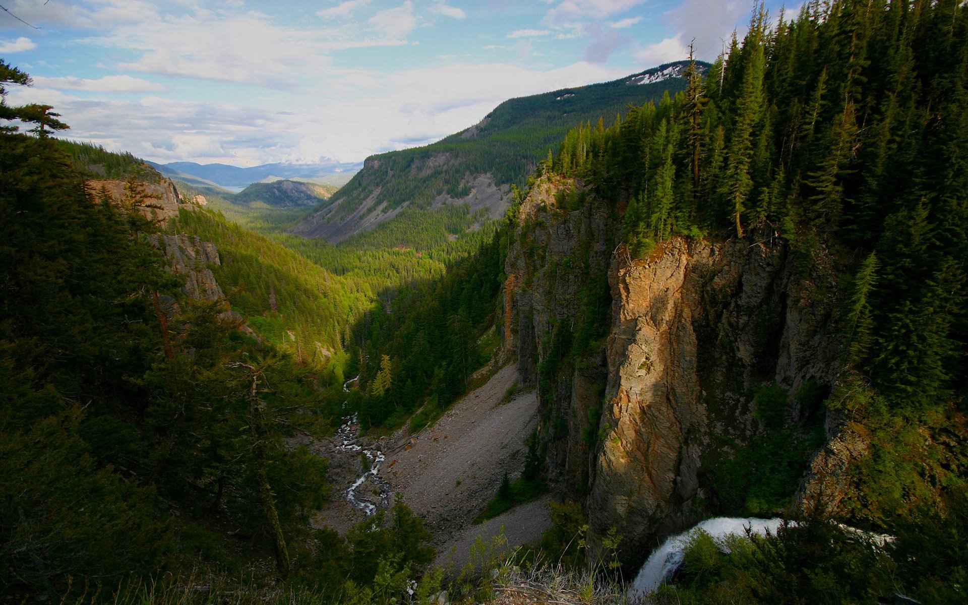 HD landscape wallpaper showcasing a lush green forest valley with a flowing stream, tall trees, and rugged mountains under a partly cloudy sky.
