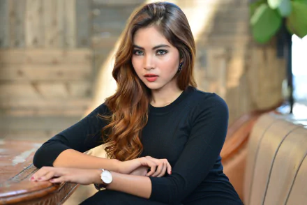 HD PC wallpaper featuring an Indonesian brunette model with striking blue eyes, seated indoors with a blurred, warm-toned background.