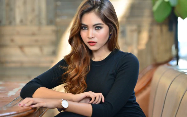 HD PC wallpaper featuring an Indonesian brunette model with striking blue eyes, seated indoors with a blurred, warm-toned background.