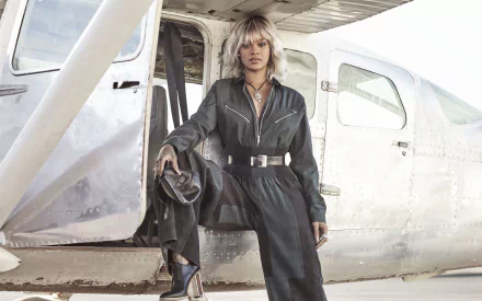 Barbadian singer Rihanna with blonde hair poses confidently in a dark jumpsuit beside a vintage airplane, captured in an HD desktop wallpaper and background.