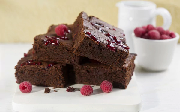 Close-up of rich chocolate brownies topped with raspberries, arranged on a white surface with a blurred background, showcased as an HD PC desktop wallpaper.