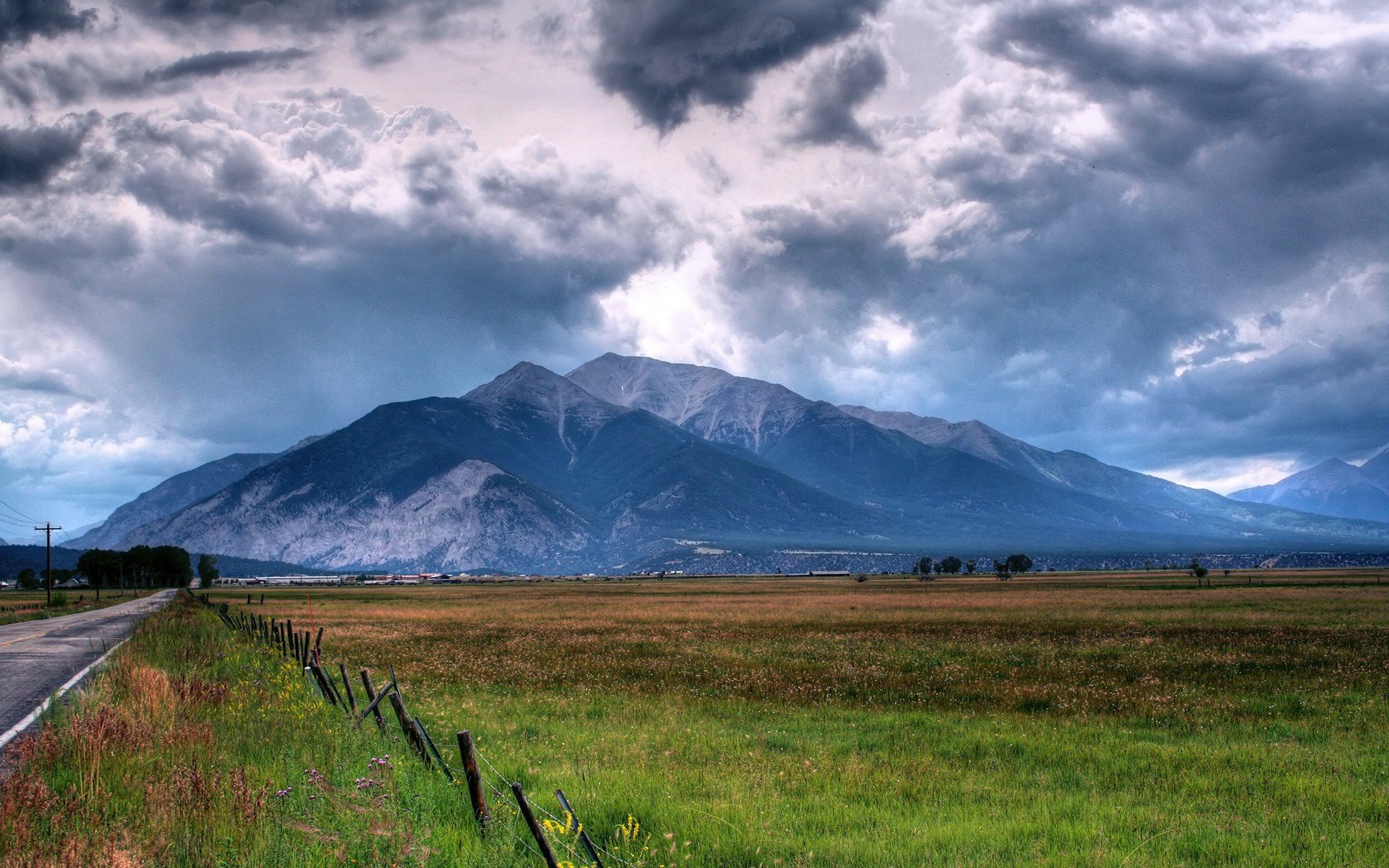 HD PC desktop wallpaper showing a vast field with a fence along a road, dramatic clouds above, and mountains in the background capturing stunning nature and landscape photography.