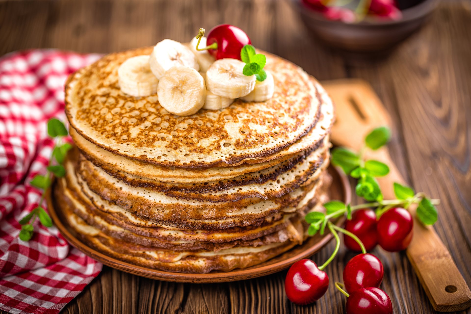 Stack of golden crêpes topped with banana slices, cherries, and a mint leaf, set on a wooden table with cherries and a red checkered cloth in a 4K Ultra HD desktop wallpaper.