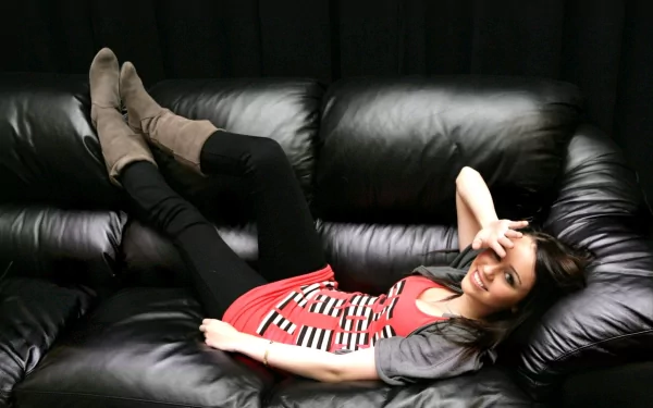 A cute, smiling Miley Cyrus lounges comfortably on a black leather couch, her playful pose and stylish outfit creating a lively and inviting HD desktop wallpaper.